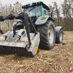 Zielony traktor z frezarką do pni drzew na polu po wycince, widoczne ścięte pnie drzew w tle.