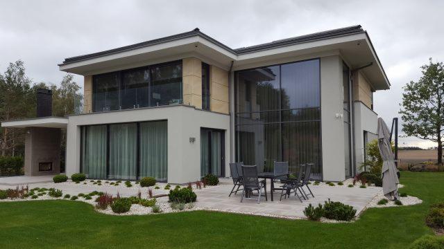 Nowoczesny dom z dużymi przeszkleniami i aluminiowymi ramami, taras z meblami ogrodowymi, uporządkowany ogród z białym żwirem.