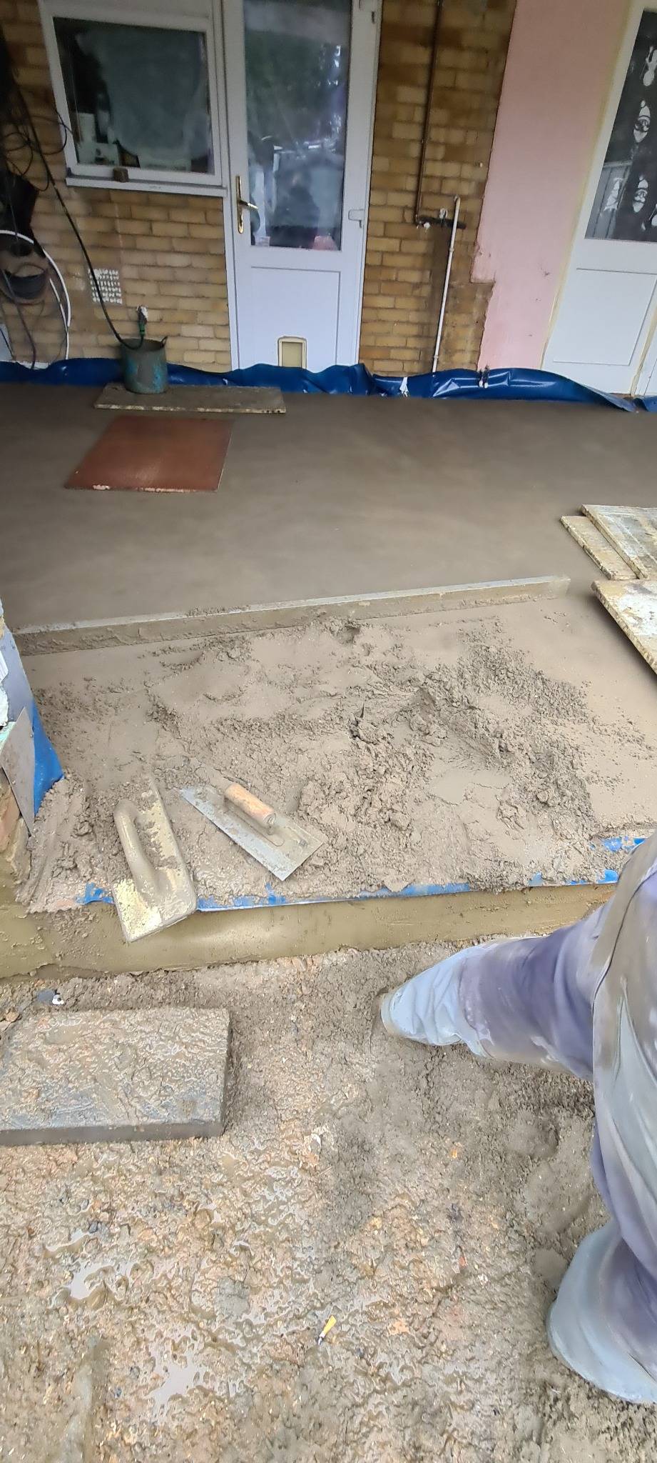 Układanie mokrego jastrychu na zewnątrz budynku, widoczne narzędzia i fragment pracownika w kombinezonie.