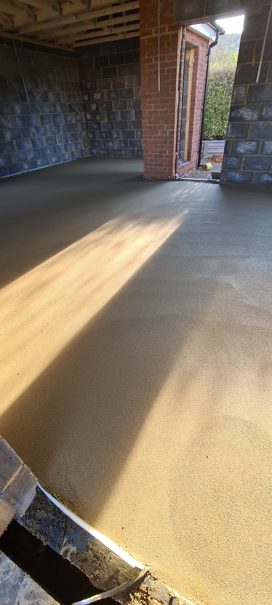 Świeżo wylana, gładka posadzka w surowym wnętrzu budynku z widocznymi ceglanymi ścianami i wiązką światła słonecznego padającą na powierzchnię.