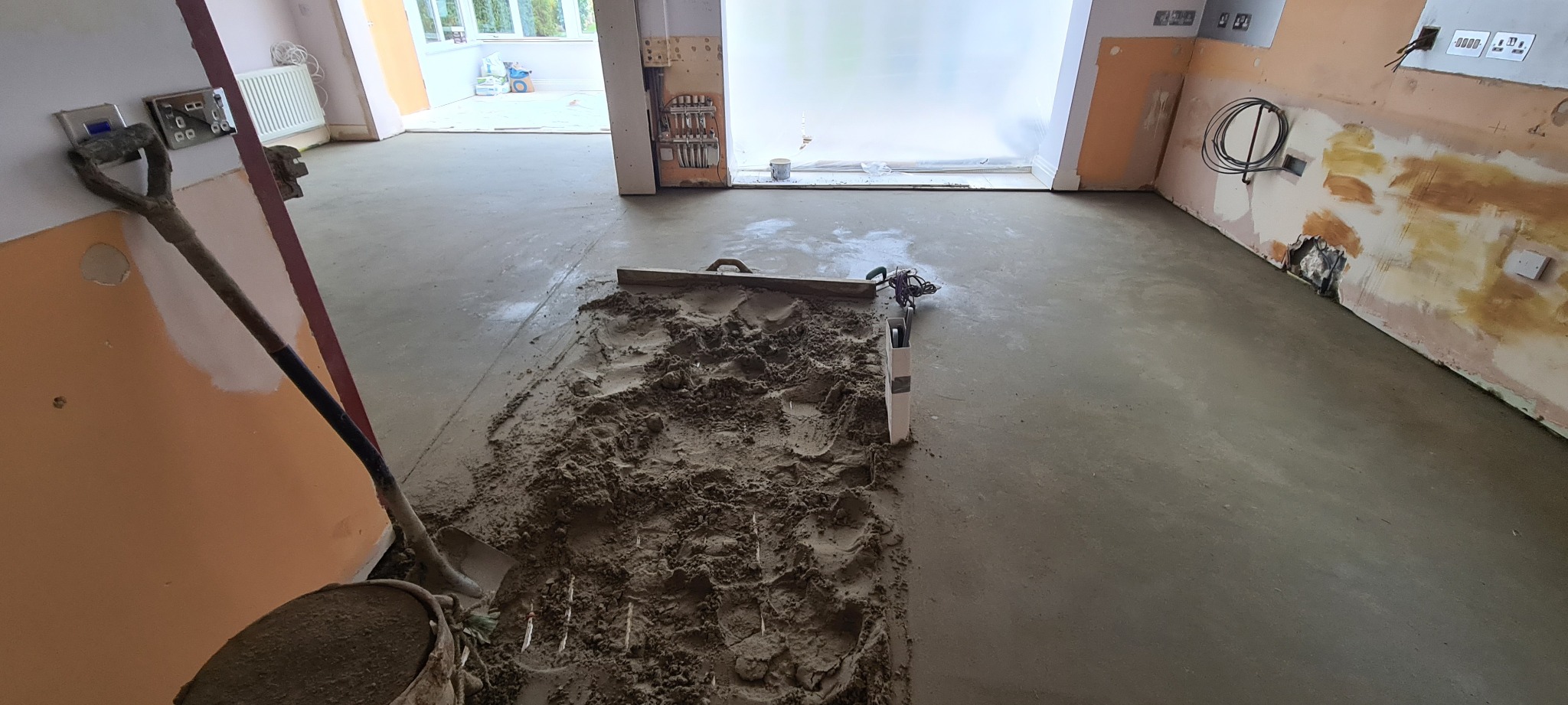 Wnętrze w trakcie wylewania posadzki, widoczne narzędzia: łopata, poziomica, listwa wyrównująca, oraz wiadro z zaprawą.