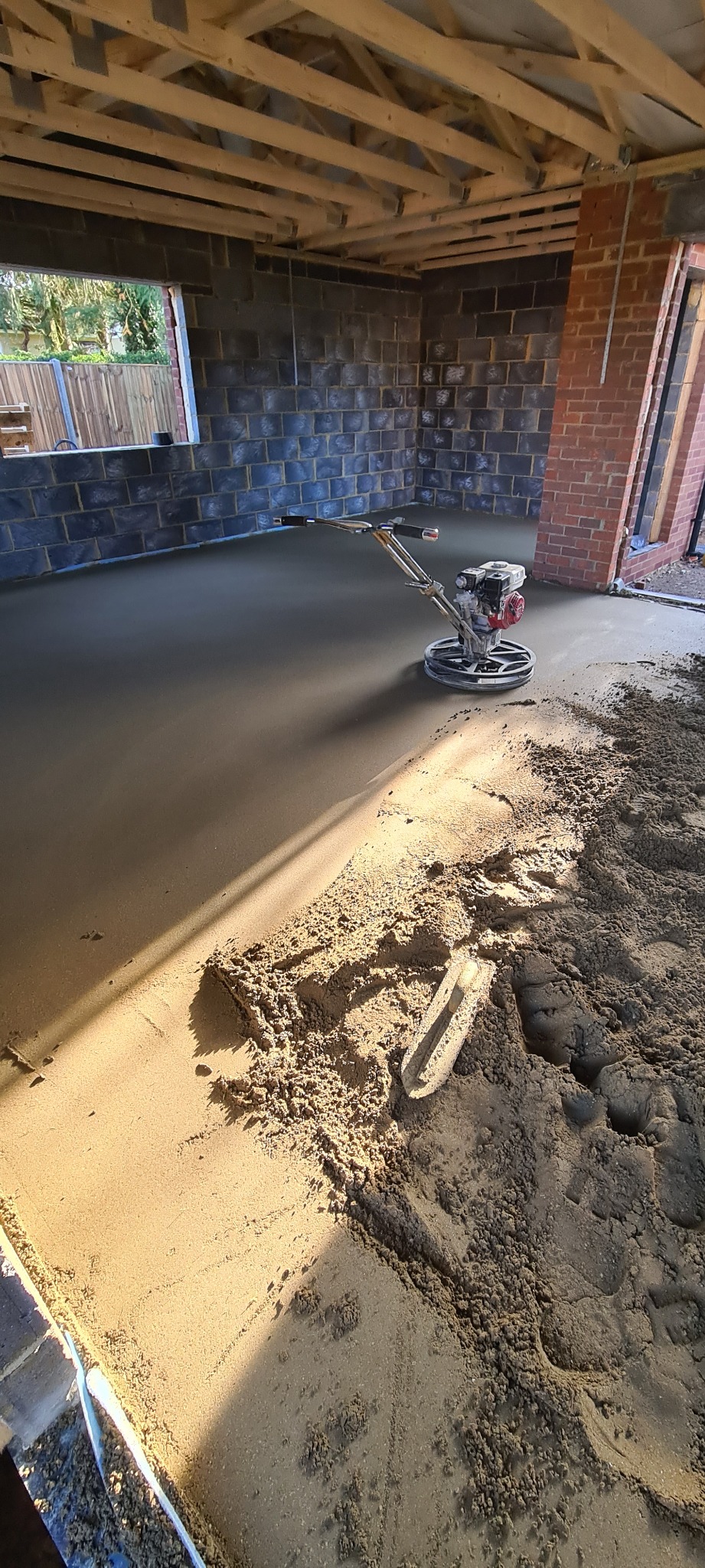 Wnętrze budynku w trakcie wylewania posadzki jastrychowej, widoczna zacieraczka mechaniczna i pryzma mokrego piasku z kielnią.