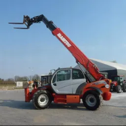 Czerwono-biały podnośnik teleskopowy Manitou MT-X 1440 z wysuniętym ramieniem i widłami, stojący na betonowym placu, w tle budynek magazynowy i czerwony traktor.