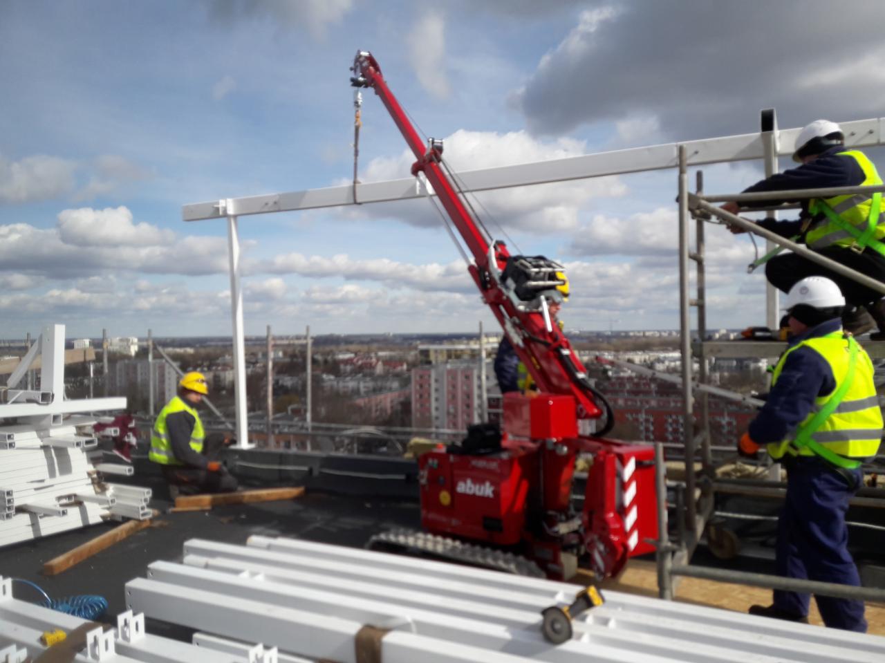 Montaż konstrukcji stalowej na dachu budynku z użyciem czerwonego mini-dźwigu gąsienicowego Abuk, pracownicy w kaskach i kamizelkach odblaskowych na tle panoramy miasta.