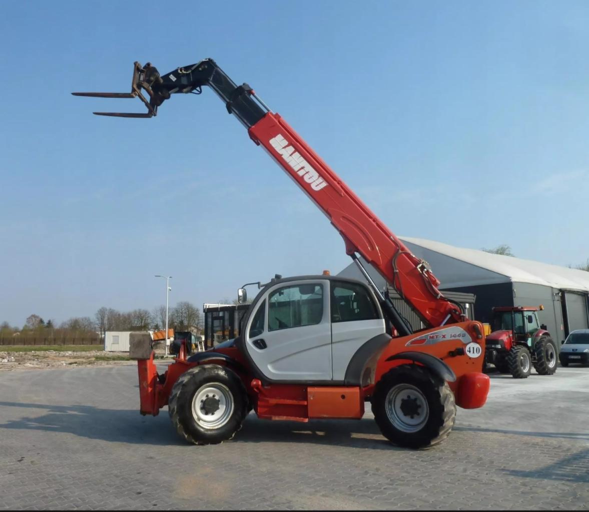 Czerwono-biały podnośnik teleskopowy Manitou MT-X 1440 z wysuniętym ramieniem i widłami, stojący na betonowym placu, w tle budynek magazynowy i czerwony traktor.