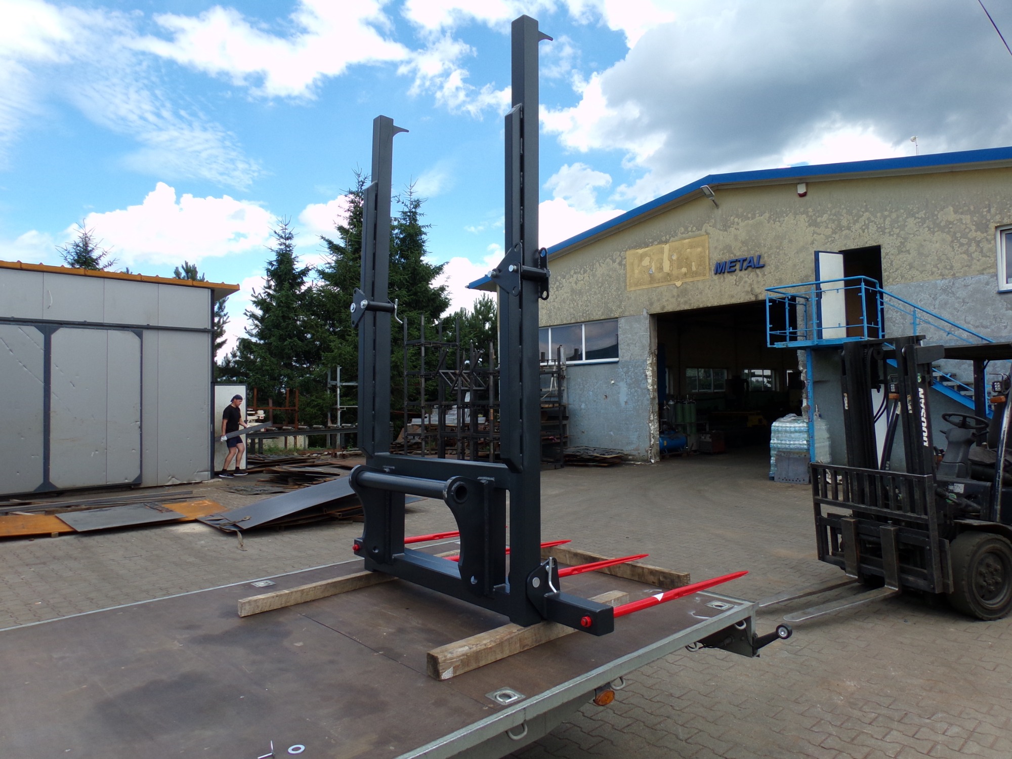 Czarna rama do bel słomy lub siana z czerwonymi widłami, umieszczona na przyczepie, w tle budynek warsztatu z napisem METAL i wózkiem widłowym.