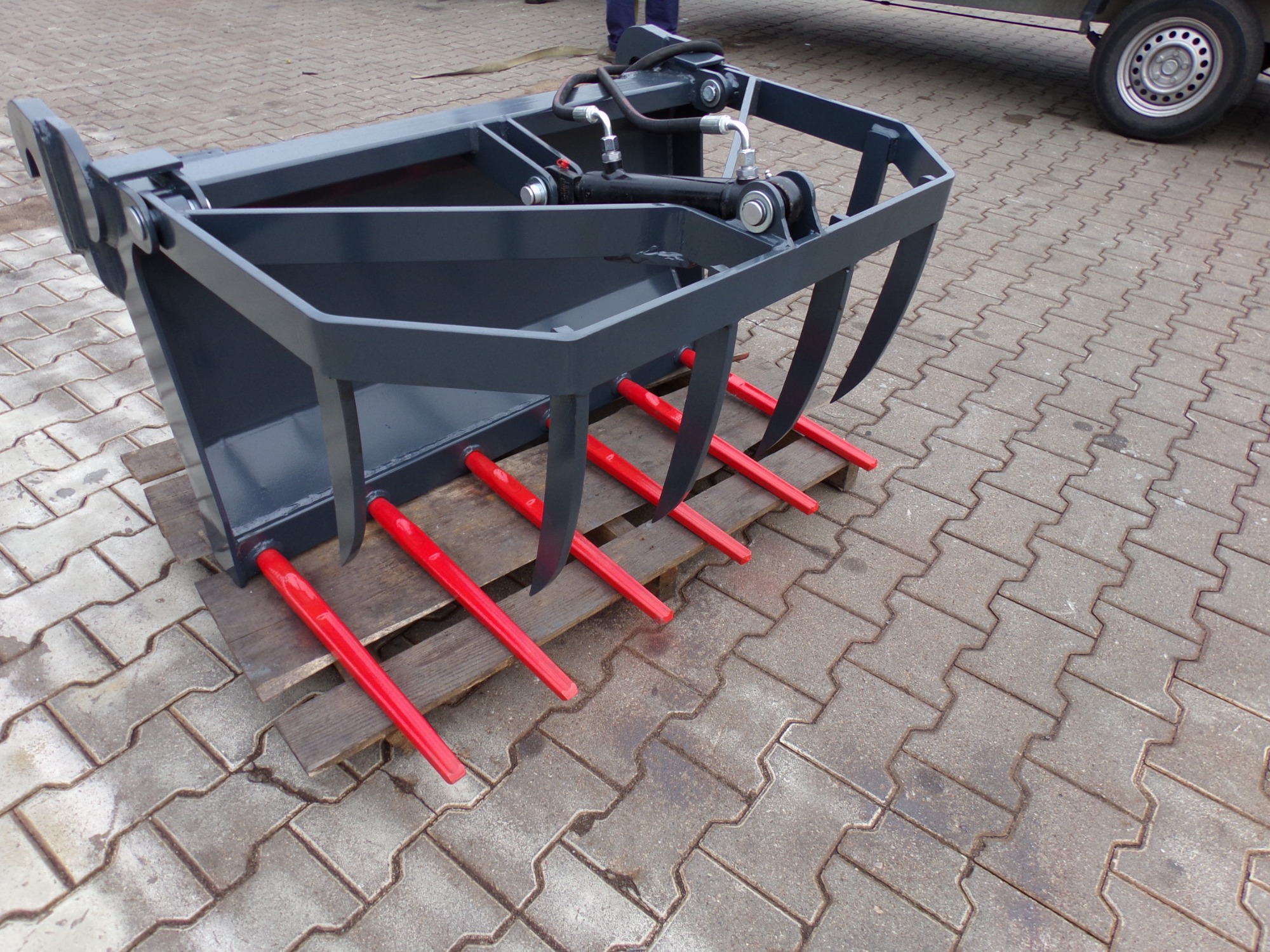 Szaro-czerwony widło-krokodyl rolniczy do ładowania obornika, leżący na palecie na kostce brukowej. Widoczne elementy hydrauliczne i mocowania.