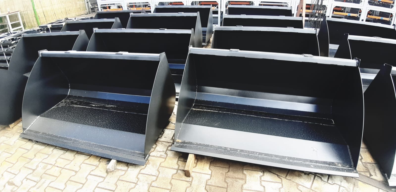 Seria nowych, czarnych łyżek do maszyn budowlanych, ułożonych na brukowanej powierzchni z widocznymi elementami konstrukcji w tle.