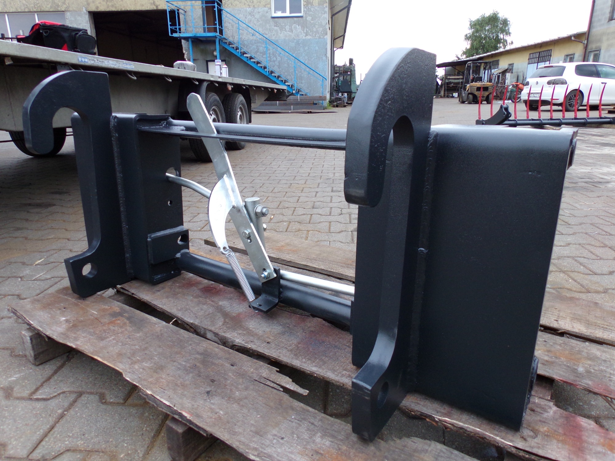 Czarny adapter ze złączem EURO, umożliwiający montaż osprzętu do koparki lub ładowarki, leżący na drewnianej palecie na zewnątrz.