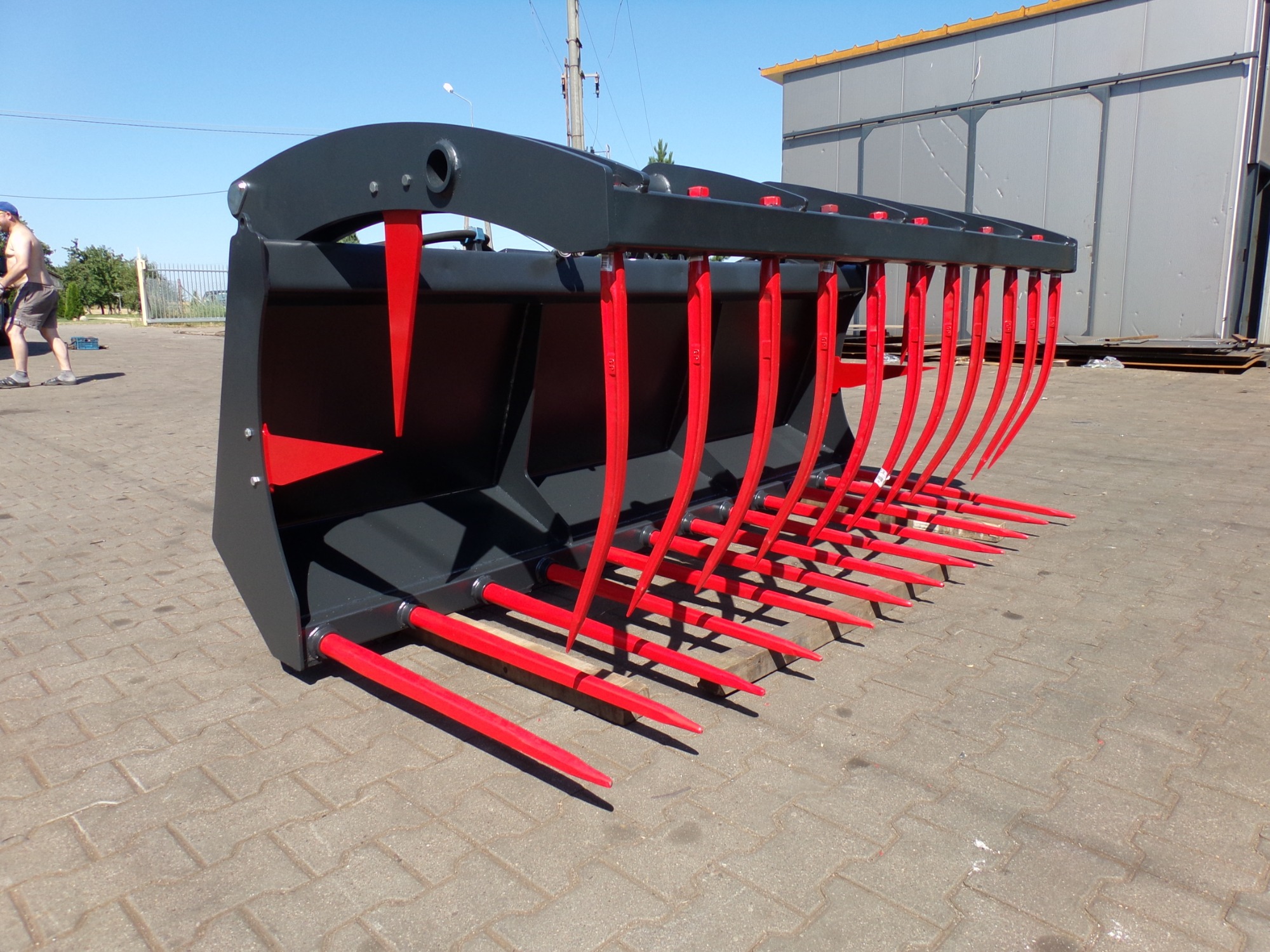 Widło-krokodyl do siana lub słomy, pomalowany na czarno z czerwonymi zębami, leżący na kostce brukowej przed szarą halą przemysłową w słoneczny dzień.