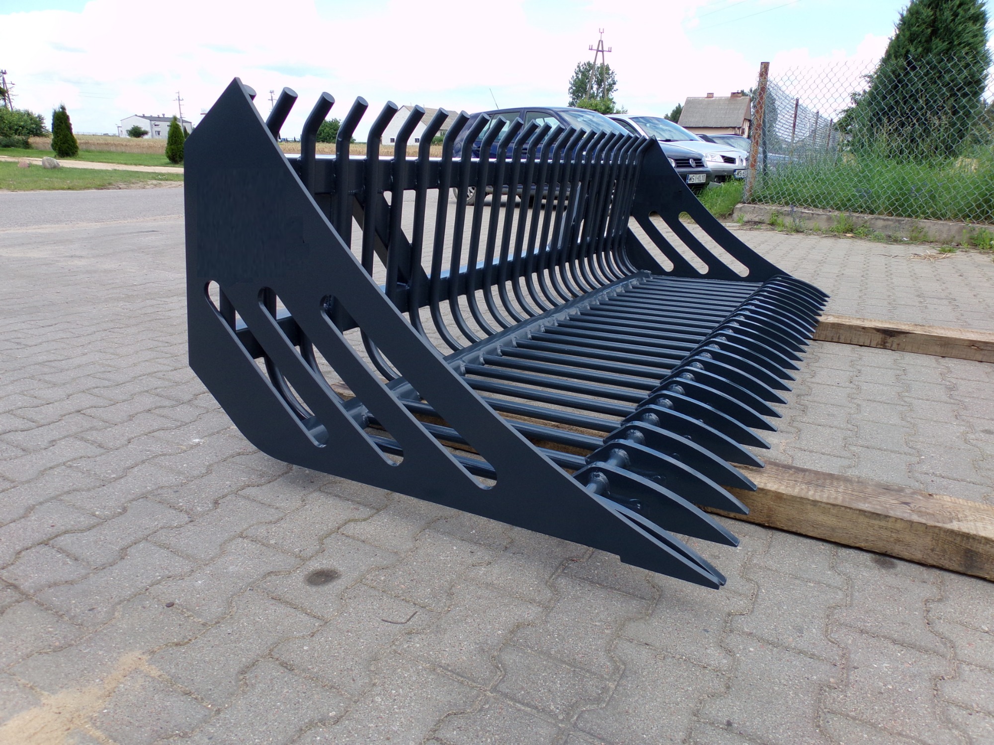 Ażurowa, pomalowana na ciemno szaro, łyżka do ładowarki lub koparki, przeznaczona do kamieni, leży na drewnianych legarach na kostce brukowej, w tle ogrodzenie z siatki i zaparkowane samochody.