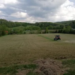 Czerwony traktor kosi trawę na rozległym, pofałdowanym polu w otoczeniu drzew i pagórków, widoczne ślady po koszeniu i pył unoszący się za maszyną.