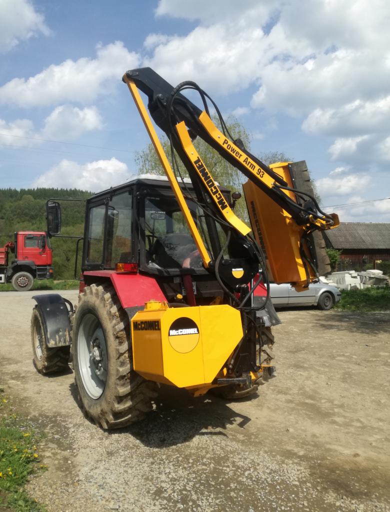 Czerwony traktor McConnell Power Arm 93 z żółtym ramieniem roboczym i głowicą tnącą, stojący na żwirowej drodze w słoneczny dzień.