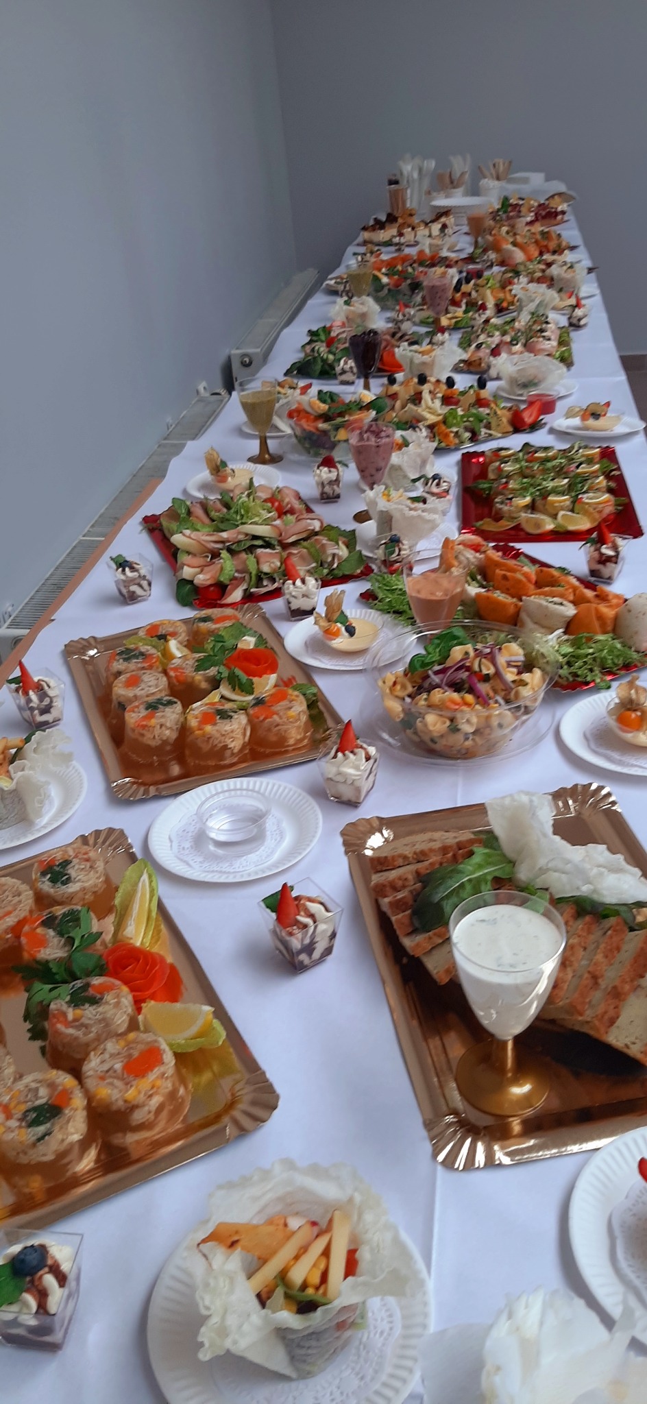 Długi stół pokryty białym obrusem, zastawiony różnorodnymi przekąskami, sałatkami i deserami w szklanych naczyniach, na złotych tacach i białych talerzach, z bliska widoczne galaretki z warzywami...