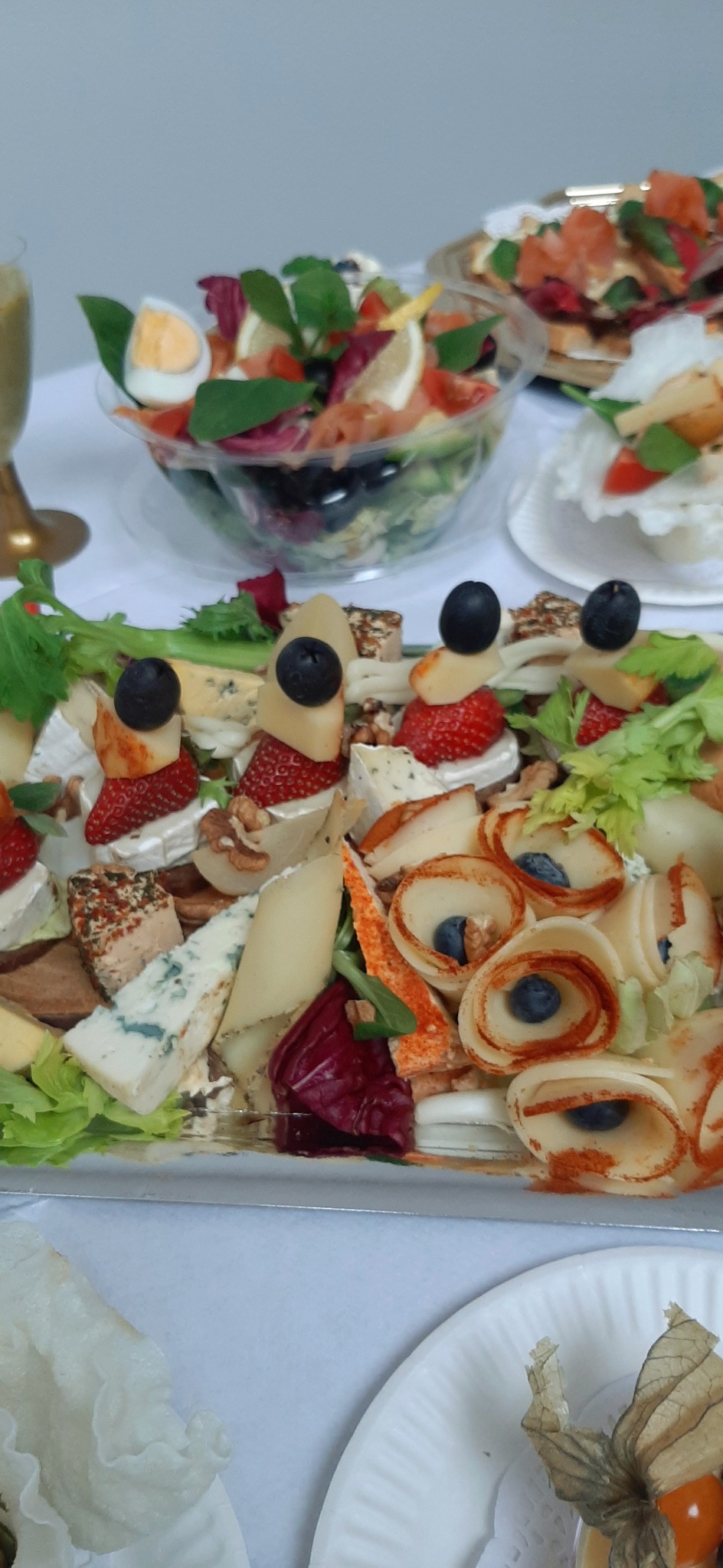 Deska serów z różnymi rodzajami sera, truskawkami, borówkami i orzechami włoskimi na srebrnej tacy, obok sałatka w przezroczystej misce i przystawki na białych talerzykach.