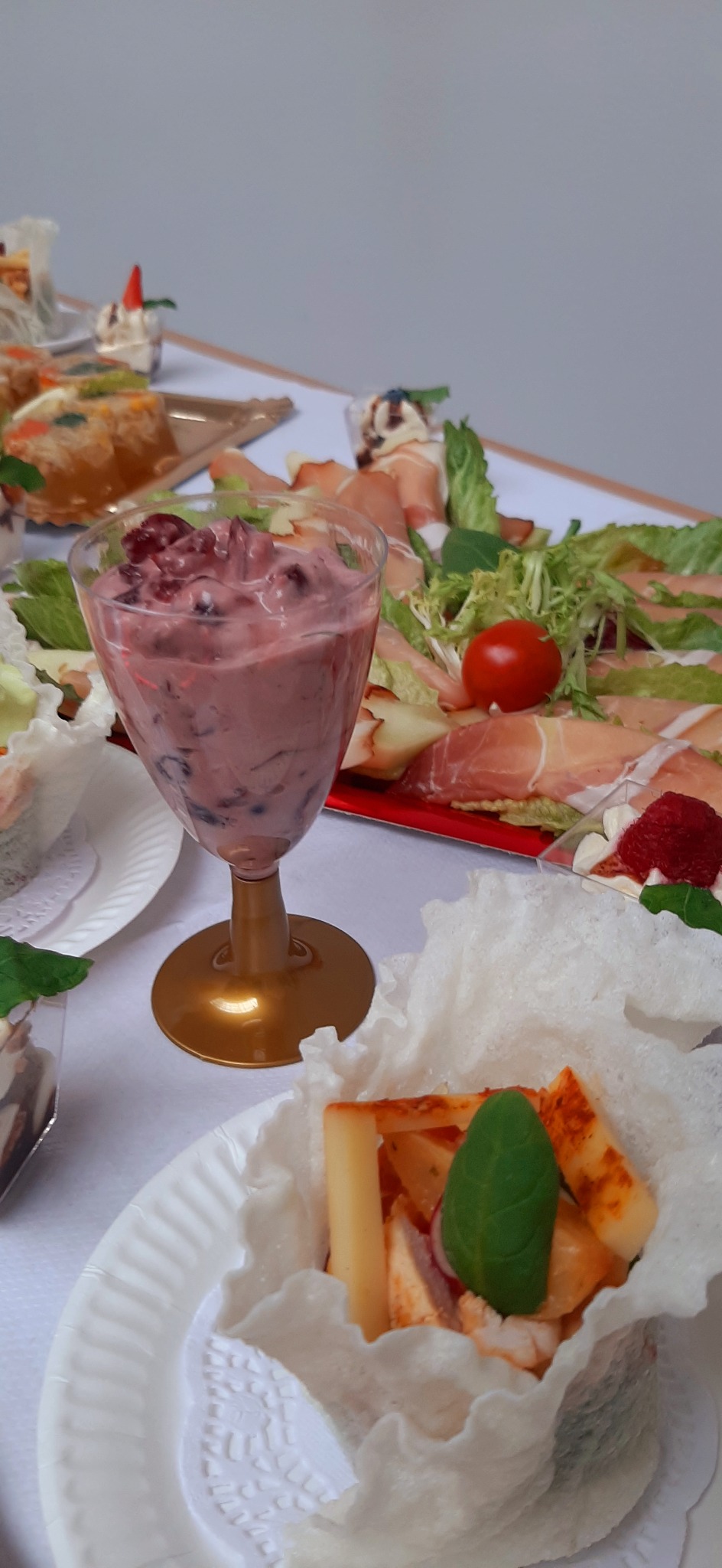 Szklanka z deserem jagodowym na złotym kielichu, obok talerz z szynką parmeńską, sałatą i pomidorem, oraz przekąska z serem i ziołami w jadalnym koszyczku na białym obrusie.