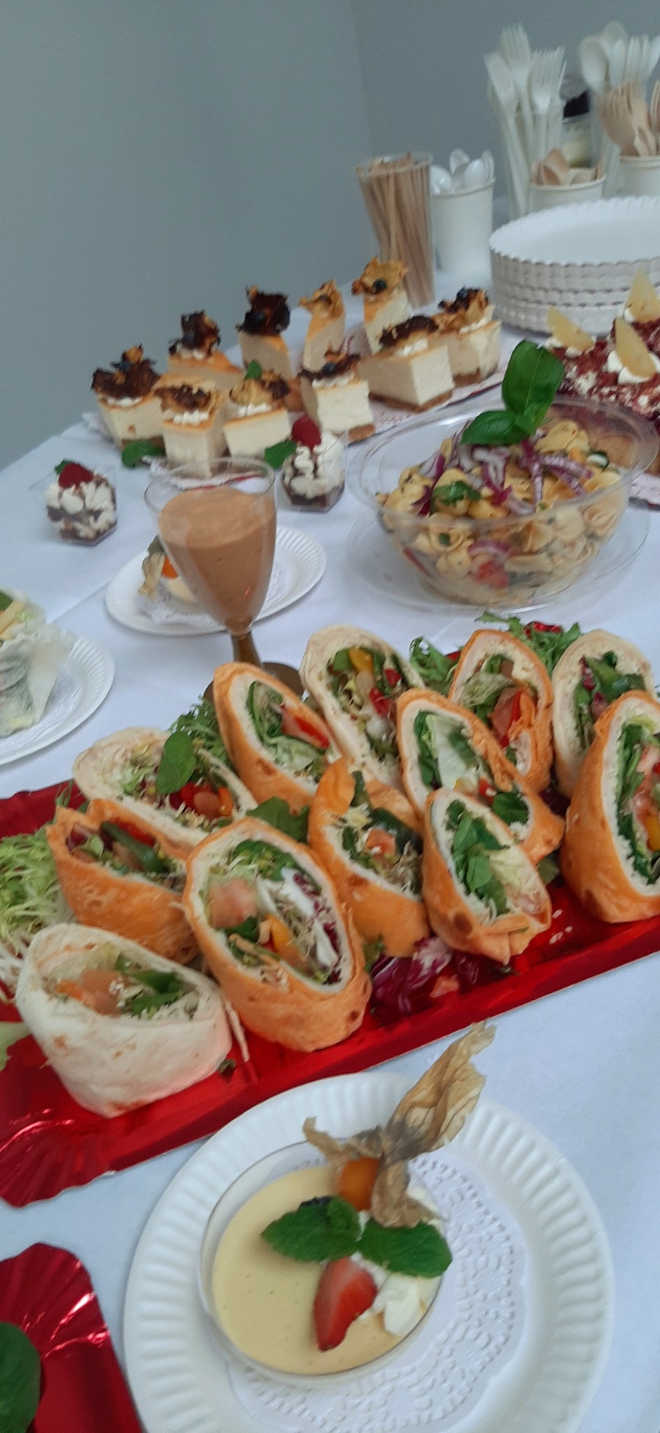 Bufet z roladkami tortilli z warzywami, tortami serowymi, sałatką makaronową i deserami w małych miseczkach, ustawiony na białym stole z plastikowymi sztućcami w tle.