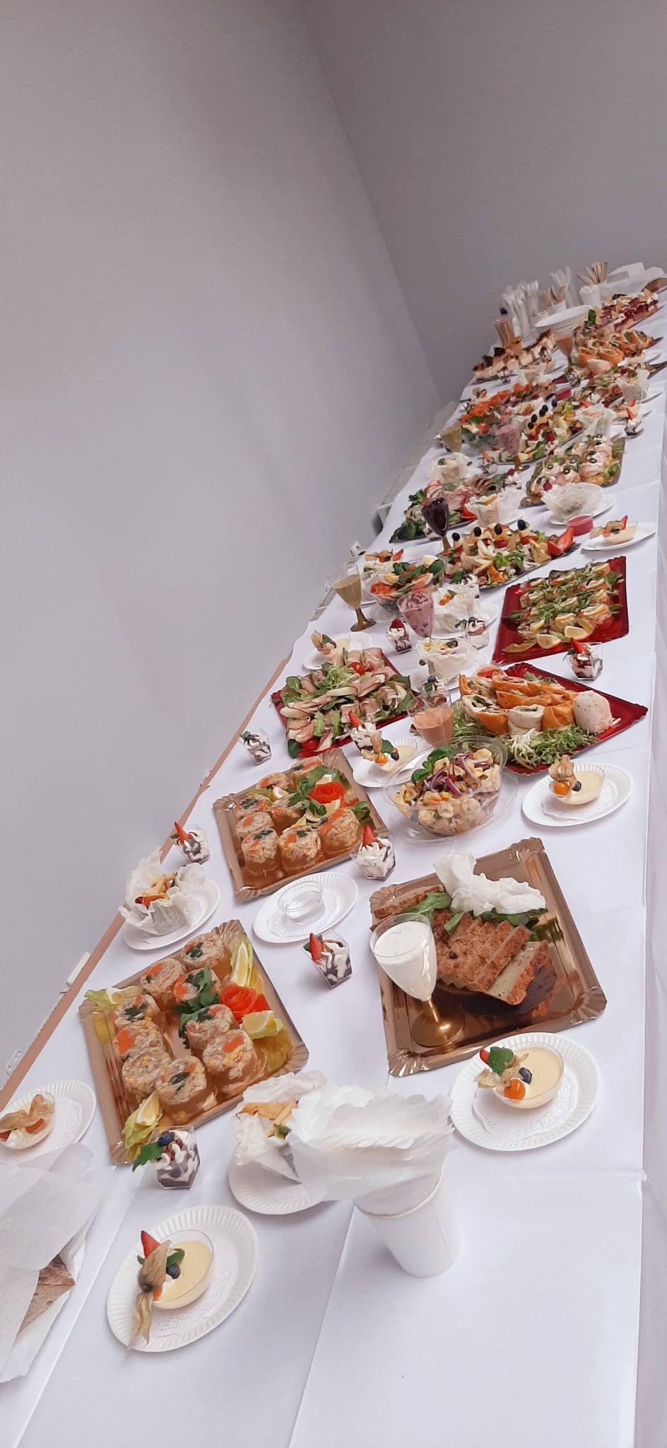 Długi stół pokryty białym obrusem, zastawiony różnorodnymi przekąskami i deserami na złotych tacach i białych talerzykach, widok z góry pod kątem. Na pierwszym planie deser w pucharku z owocami...