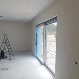 Wnętrze w trakcie prac wykończeniowych: ściany pokryte gładzią, widoczna drabina malarska, przesuwne okno zabezpieczone folią, przygotowane punkty oświetleniowe na suficie.