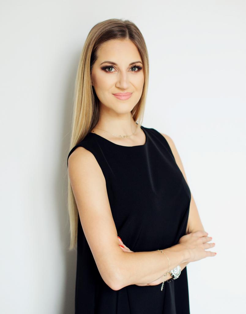 Portret kobiety z długimi blond włosami w czarnej sukience, z rękami skrzyżowanymi, na tle białej ściany, z delikatnym uśmiechem i subtelnym makijażem, z widoczną bransoletką i zegarkiem.