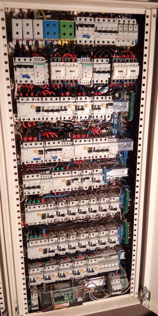 Rozdzielnica elektryczna z licznymi bezpiecznikami, przekaźnikami i okablowaniem w metalowej obudowie z perforowanymi bokami, widoczne różne kolory przewodów i zacisków.