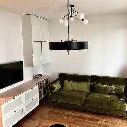 Nagel Meble - Nowoczesny salon z zieloną sofą, szafką RTV w kolorze brzoskwini i białymi szafkami wiszącymi, oświetlony designerską lampą wiszącą z kulistymi kloszami.