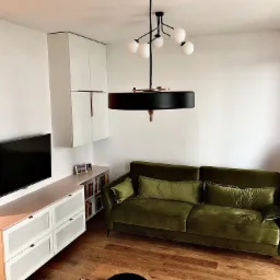 Nowoczesny salon z zieloną sofą, szafką RTV w kolorze brzoskwini i białymi szafkami wiszącymi, oświetlony designerską lampą wiszącą z kulistymi kloszami.