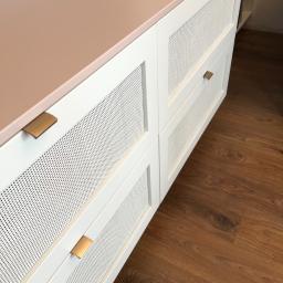 Nagel Meble - Biała komoda z ażurowymi frontami szuflad i blatem w kolorze pudrowego różu, z minimalistycznymi, metalowymi uchwytami, na tle drewnianej podłogi.