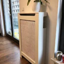 Biała obudowa grzejnika z plecionką, na niej doniczka z zieloną rośliną, w tle okno z widokiem na budynek.