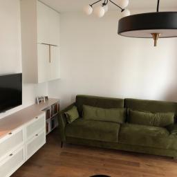 Nagel Meble - Salon z zieloną sofą, szafką RTV z białymi frontami z rattanem, białymi szafkami wiszącymi z złotymi uchwytami i drewnianą podłogą.