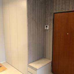 Nagel Meble - Wnętrze przedpokoju z dużą, jasną szafą w zabudowie, obok wejścia z brązowymi drzwiami, ściana wykończona szarymi panelami tapicerowanymi, mała szafka z siedziskiem.