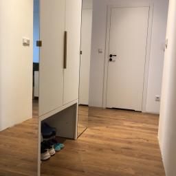 Nagel Meble - Wnętrze przedpokoju z lustrzanym panelem szafy, drewnianą podłogą i białymi drzwiami z czarną klamką.