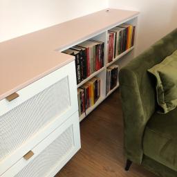 Nagel Meble - Biała szafka z ażurowymi frontami szuflad, różowy blat, wbudowany regał na książki, widok z boku zielonej sofy.