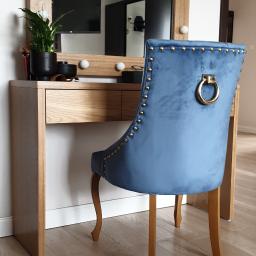 Nagel Meble - Toaletka z lustrem otoczonym żarówkami i aksamitnym krzesłem w kolorze niebieskim, widok z tyłu na ozdobne nity i złoty uchwyt na oparciu.