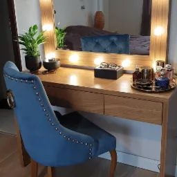 Toaletka z lustrem otoczonym żarówkami, drewniany blat, niebieskie krzesło z ozdobnymi nitami, akcesoria do makijażu na blacie, roślina doniczkowa, widok na sypialnię w odbiciu lustra.