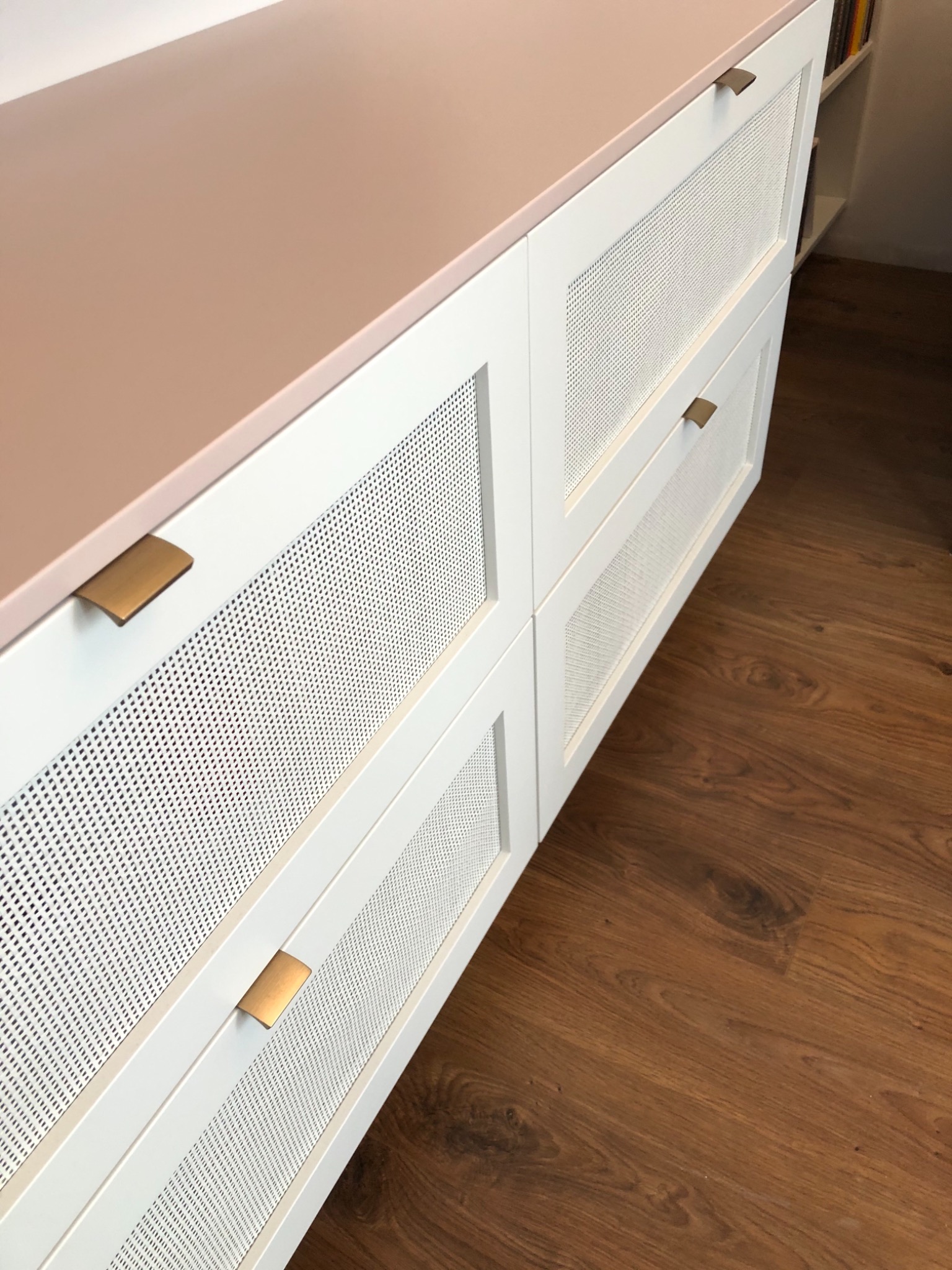 Biała komoda z ażurowymi frontami szuflad i blatem w kolorze pudrowego różu, z minimalistycznymi, metalowymi uchwytami, na tle drewnianej podłogi.
