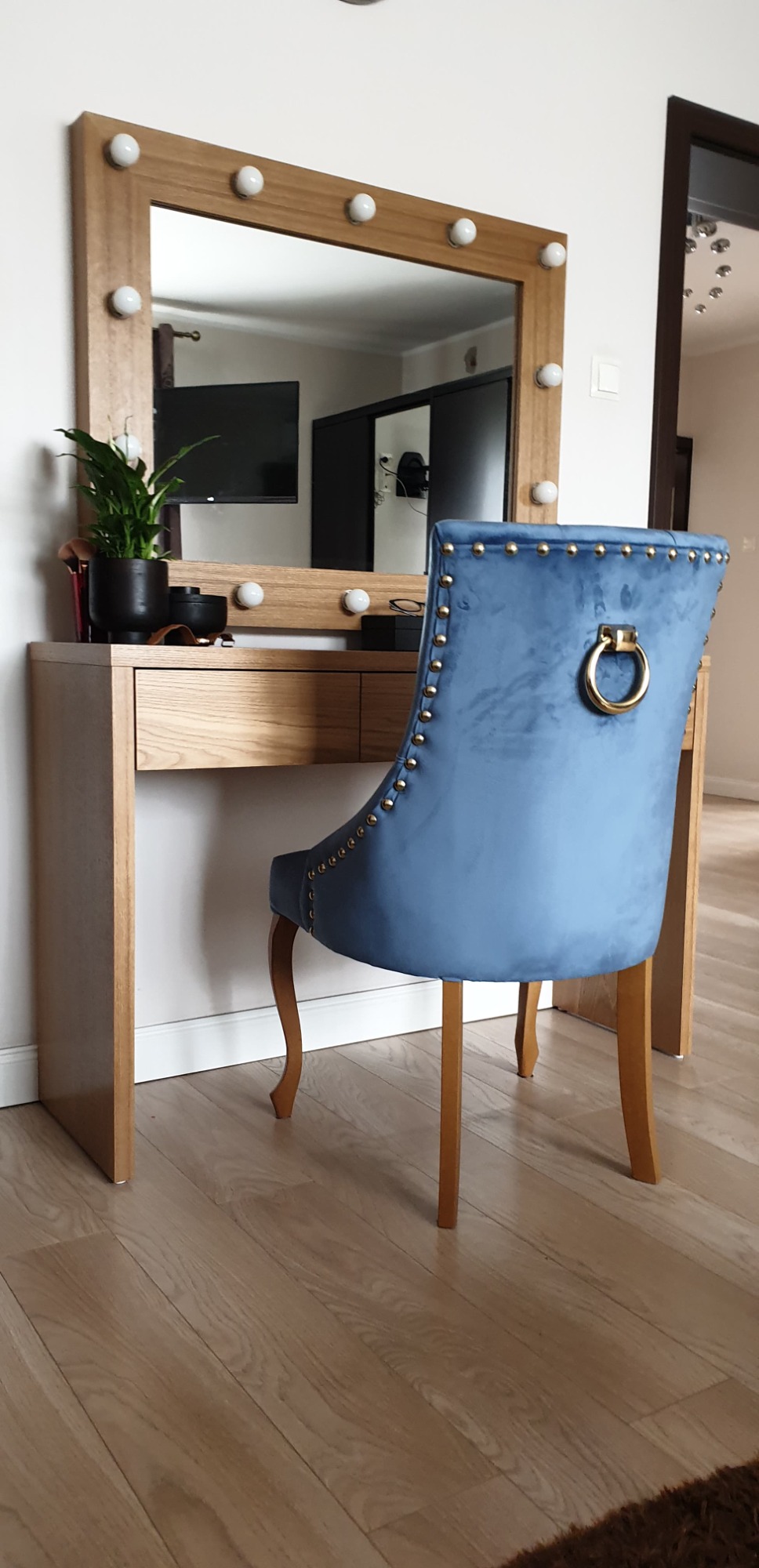 Toaletka z lustrem otoczonym żarówkami i aksamitnym krzesłem w kolorze niebieskim, widok z tyłu na ozdobne nity i złoty uchwyt na oparciu.