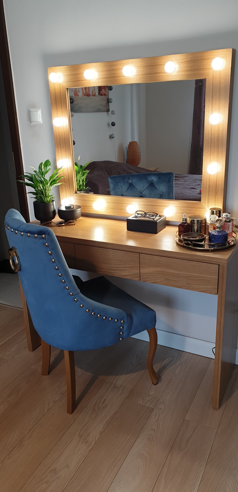 Toaletka z lustrem otoczonym żarówkami, drewniany blat, niebieskie krzesło z ozdobnymi nitami, akcesoria do makijażu na blacie, roślina doniczkowa, widok na sypialnię w odbiciu lustra.