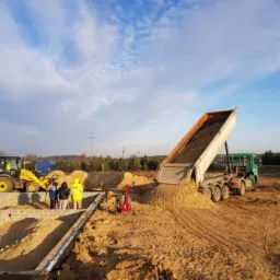 Wyładunek piasku wywrotką na placu budowy fundamentów z bloczków betonowych, widoczny mały traktor i grupa osób w odblaskowych kamizelkach.