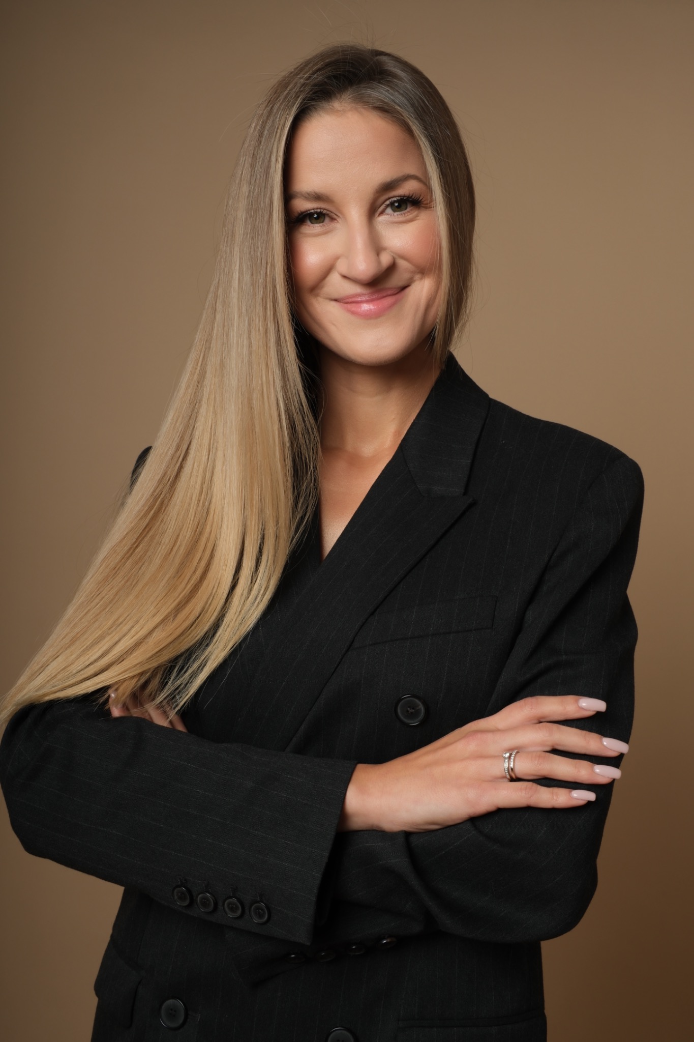 Uśmiechnięta blondynka w czarnym, eleganckim żakiecie w prążki, z założonymi rękami na beżowym tle. Widoczny pierścionek na palcu.