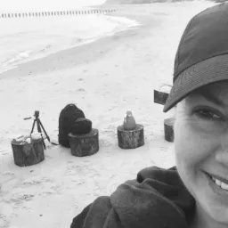 Czarno-białe zdjęcie z plaży. Na pierwszym planie uśmiechnięta kobieta w czapce. W tle sprzęt fotograficzny, plecak i termos na pniakach, oraz brzeg morza.