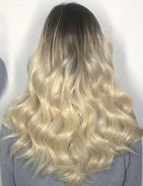 Długie, falowane włosy z ombre, ciemne u nasady przechodzące w platynowy blond, widoczne od tyłu, na tle białej ściany i szarego swetra.