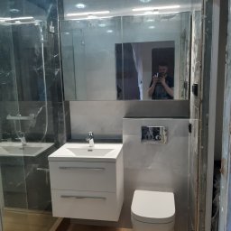 Bianko-Bud - Nowoczesna łazienka z kabiną prysznicową, umywalką z szafką i podwieszaną toaletą. Szare płytki imitujące marmur. W lustrze odbija się osoba robiąca zdjęcie.