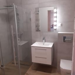 Bianko-Bud - Nowoczesna łazienka z kabiną prysznicową typu walk-in, umywalką z szafką, toaletą podwieszaną i lustrem z oświetleniem LED, wykończona szarymi płytkami ceramicznymi i drewnopodobną podłogą.