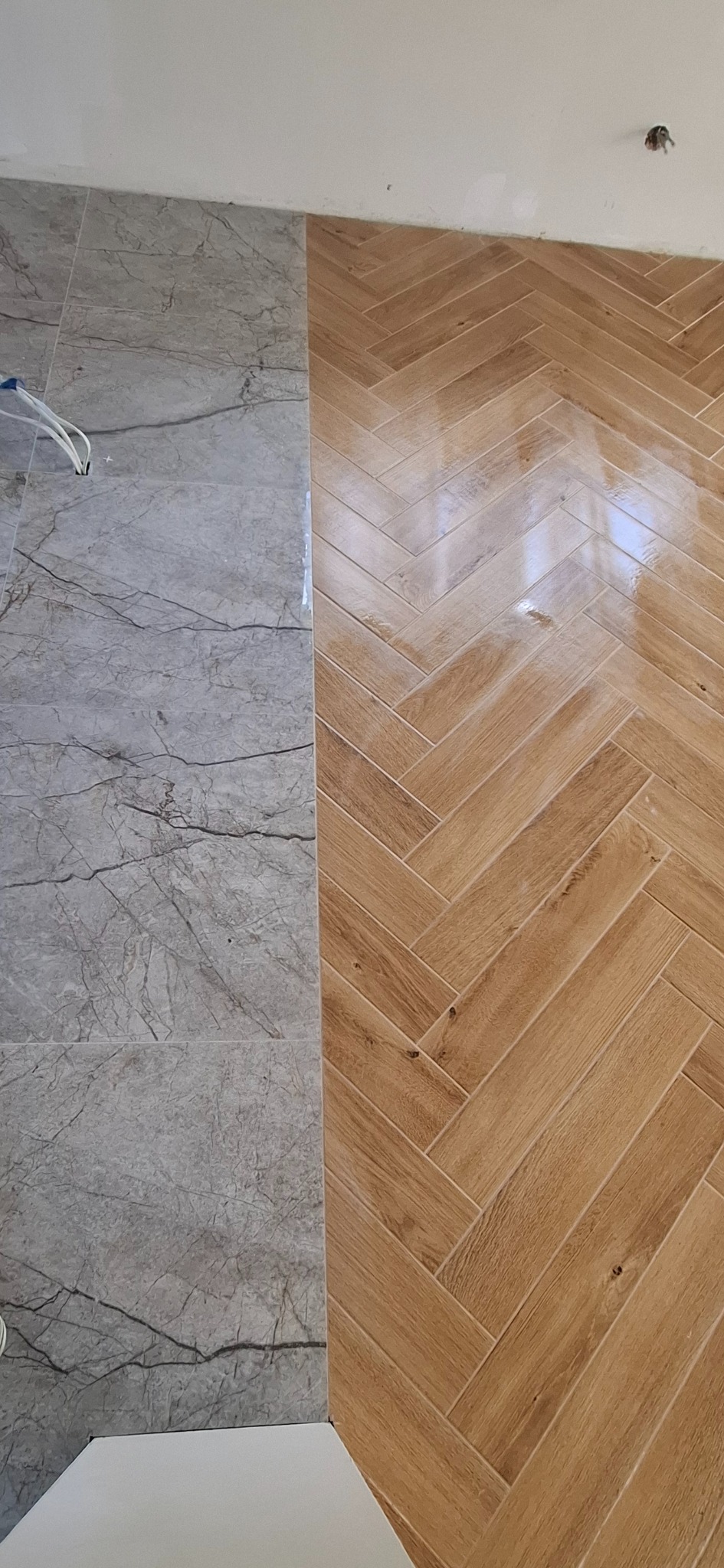 Ukończona podłoga z płytek: szare płytki imitujące marmur zestawione z drewnopodobnymi płytkami ułożonymi w jodełkę. Widoczne kable elektryczne.