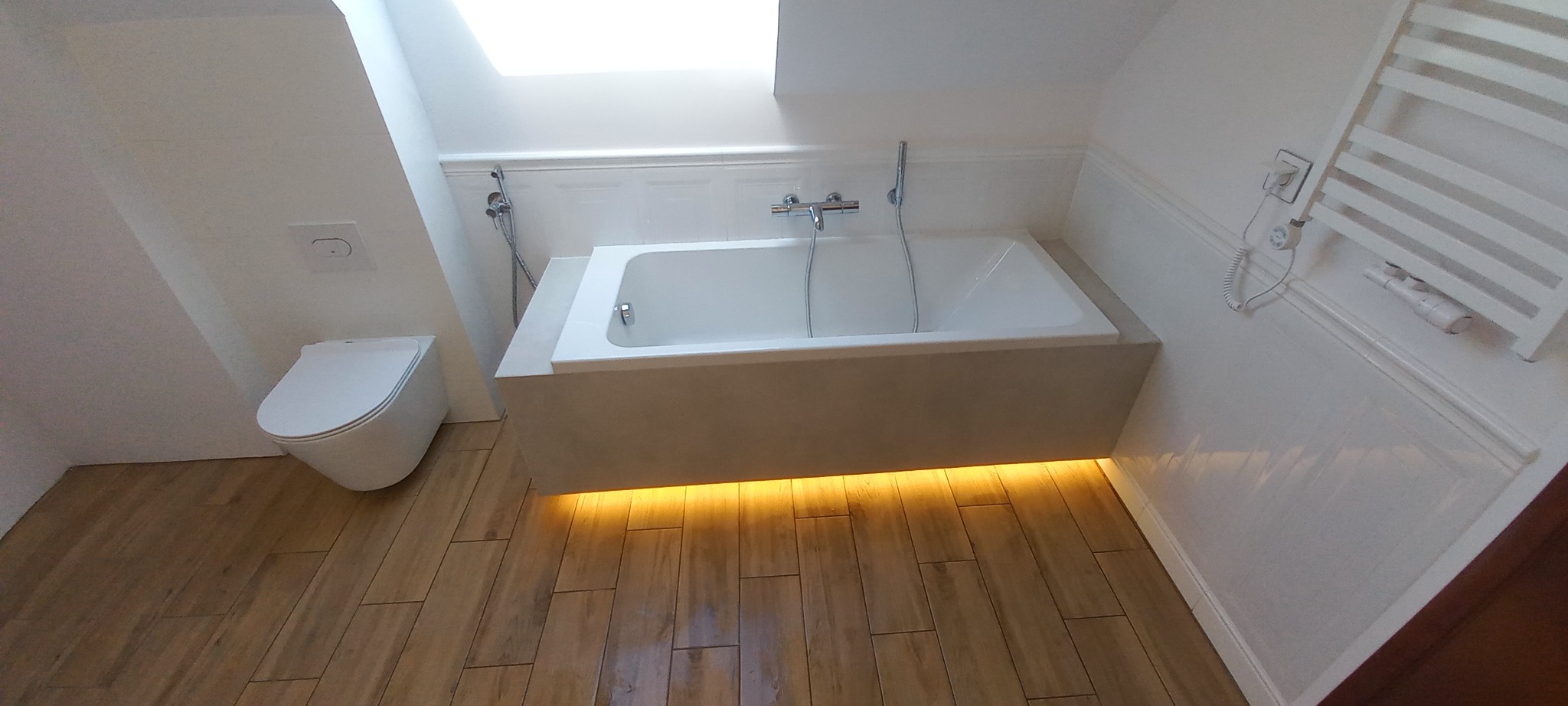 Nowoczesna łazienka na poddaszu: biała wanna z podświetleniem LED, wisząca toaleta i grzejnik drabinkowy na tle białych ścian i drewnopodobnej podłogi.