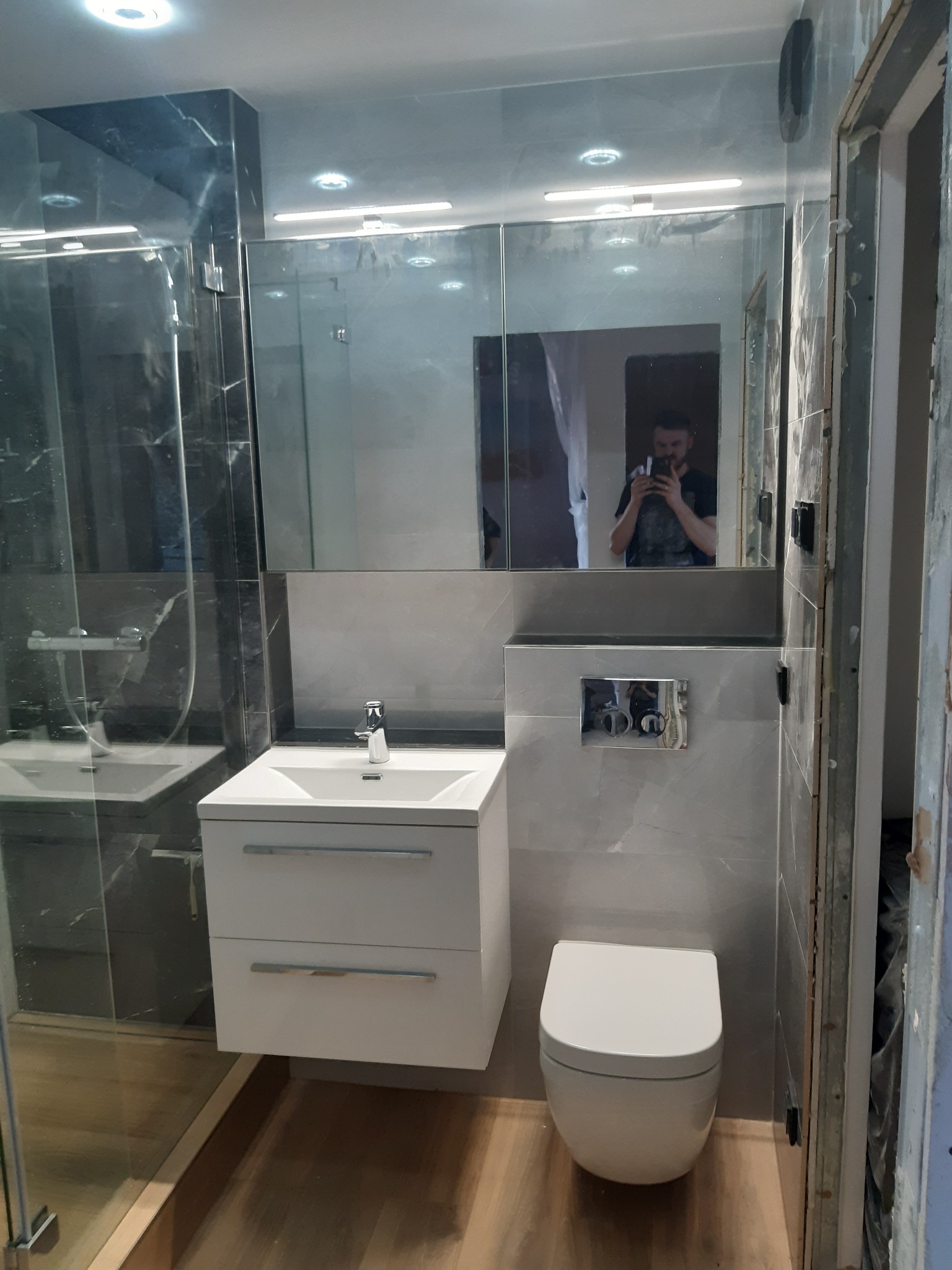 Nowoczesna łazienka z kabiną prysznicową, umywalką z szafką i podwieszaną toaletą. Szare płytki imitujące marmur. W lustrze odbija się osoba robiąca zdjęcie.
