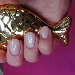 Dłoń z delikatnym, błyszczącym manicure hybrydowym, jeden paznokieć ozdobiony srebrnym wzorem, trzymająca pędzel do makijażu o złotym uchwycie w kształcie ryby na tle fuksjowego materiału.