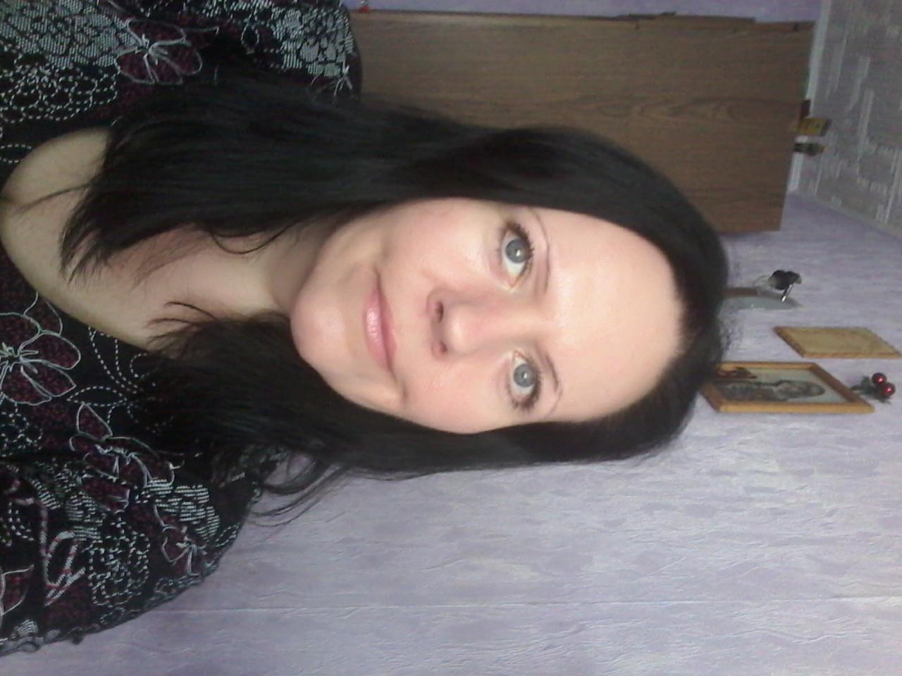 Portret kobiety z ciemnymi włosami i jasnymi oczami, lekko uśmiechniętej, na tle ściany i drewnianego mebla. Ujęcie z bliska.