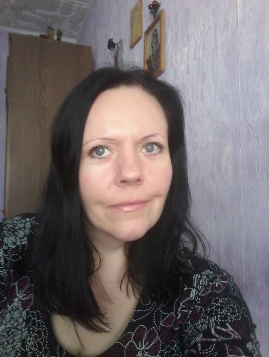 Portret kobiety z ciemnymi włosami na tle fioletowej ściany. W tle widoczny fragment szafy i obrazek. Osoba patrzy wprost, uśmiechając się delikatnie.